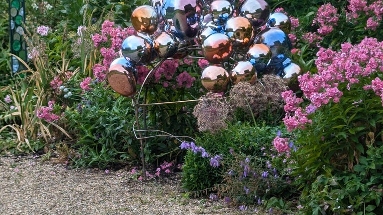 A sculpture of colourful metal bubbles installed in a flower bed, with pink and purple flowers and shrubs surrounding it.